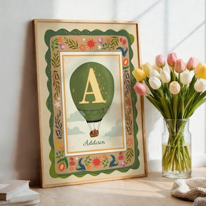 May include: A framed rectangular wall art print featuring a green hot air balloon with a large gold letter A in the centre. Below the balloon, the name Addison is printed. The border is decorated with colourful flowers, suns, and stars.