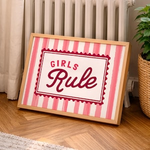 May include: A framed sign with a light wood frame. The sign features a white background with pink vertical stripes. The centre of the sign has a white rectangle with the words "GIRLS Rule" in a burgundy script font.