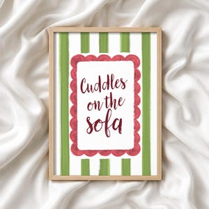May include: A framed print with the words "Cuddles on the sofa" in a decorative font. The background features green vertical stripes on a white backdrop. The text is inside a red scalloped border.
