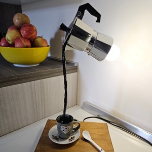 Può includere: Una base in legno con una tazza e un piattino in ceramica bianca, un cucchiaio bianco e una caffettiera argentata con una lampadina all'interno che versa liquido nero nella tazza.