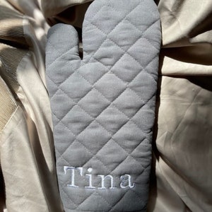 May include: Grey quilted oven mitt with the name "Tina" embroidered in white.