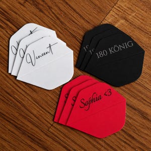 Könnte beinhalten: Drei Sätze Dart-Flights in Weiß, Schwarz und Rot. Die weißen Flights tragen den Namen Vincent in schwarzer Schrift. Die schwarzen Flights sind mit 180 KÖNIG in Weiß bedruckt. Die roten Flights zeigen den Namen Sophia <3 in Schwarz.