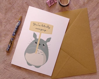 May include: A white card with a gray cartoon character holding a sign that says "You're totally amazing". The card is on a pink fuzzy surface with a brown envelope and a pink paperclip.