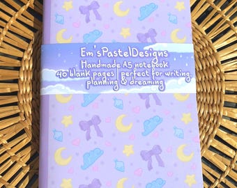 May include: A lavender A5 notebook with a repeating pattern of moons, stars, bows, and clouds. The notebook has a band that reads "Em's Pastel Designs Handmade A5 notebook 40 blank pages perfect for writing, planning & dreaming."