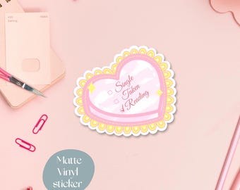 May include: A matte vinyl sticker in a heart shape with a pink and yellow design. The sticker has the text "Single, Taken, Reading". The sticker is surrounded by other stationery items on a pink surface.