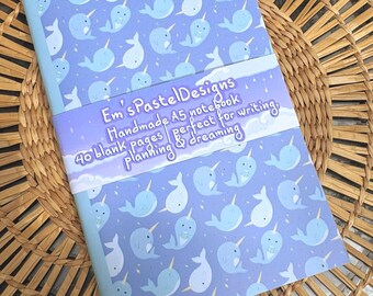 May include: A handmade A5 notebook with a light blue cover featuring a pattern of cartoon narwhals. The notebook has 40 blank pages and a band that reads "Em's Pastel Designs" and "perfect for writing, planning & dreaming."
