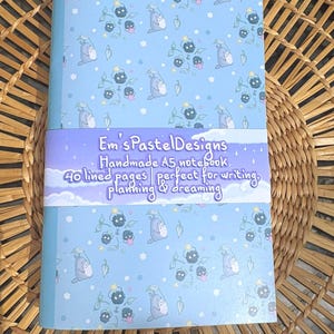 May include: A pastel blue A5 notebook featuring a pattern of cartoon characters and floral designs. The notebook has a purple band with the text "Em's Pastel Designs" and "Handmade A5 notebook, 40 lined pages perfect for writing, planning & dreaming."