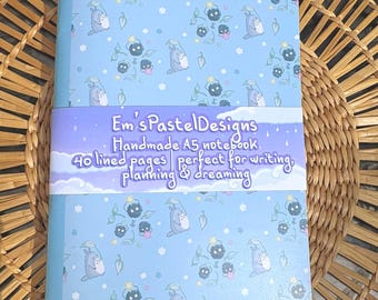 May include: A light blue A5 notebook with a repeating pattern of cartoon characters and floral elements. The notebook has a purple band with the text "Em's Pastel Designs" and "Handmade A5 notebook, 40 lined pages perfect for writing, planning & dreaming."