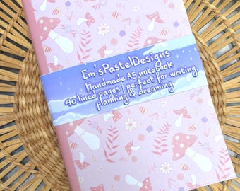 May include: A5 notebook with a pink cover featuring a pattern of mushrooms, flowers, and leaves. The notebook has a light blue band with the text "Em's Pastel Designs" and "Handmade A5 notebook perfect for writing, planning & dreaming."