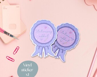 May include: Two light purple "Stationery Addict" vinyl stickers with a ribbon design. The stickers have a light purple border and feature a pencil and a small bag graphic. The image also includes pink pens, paperclips, and a pair of scissors.