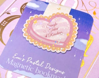 May include: A pastel purple magnetic bookmark with a heart-shaped design. The heart has a pink border and the words "Single Taker Reading". The bookmark is on a light purple book with gold floral accents. The text "Em's Pastel Designs Magnetic bookmark" is also visible.