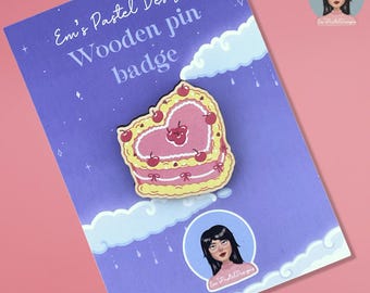 May include: A wooden pin badge in the shape of a heart-shaped cake with cherries on top. The pin is pink and yellow, with the text "Em's Pastel Designs" and "Wooden pin badge" on the backing card. The background is purple.