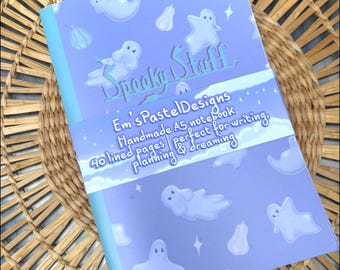 May include: A handmade A5 notebook with a light blue cover featuring ghost illustrations and the text "Spooky Stuff". The notebook has 40 lined pages and is perfect for writing, planning, and dreaming. The notebook is on a woven basket.