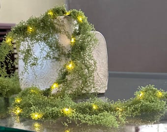 Puede incluir: Un jarrón de cerámica blanca con superficie texturizada está cubierto por una guirnalda verde similar al musgo que presenta pequeñas luces LED de color amarillo cálido. La guirnalda rodea el jarrón y se extiende, creando un adorno botánico.