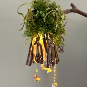May include: A decorative hanging ornament shaped like a bell, constructed from dark brown twigs arranged vertically, topped with a cluster of bright green moss, and illuminated from within by a warm yellow light, featuring small dangling beads.