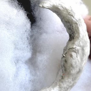 May include: A silver-colored crescent moon ornament with a sculpted face featuring closed eyes and a nose. The surface has a textured, metallic finish with visible indentations and contours, set against a background of white, fluffy material.