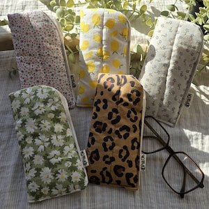Peut inclure: Plusieurs étuis à lunettes en tissu avec différents motifs : floral, citron, et léopard. Les étuis sont de couleurs rose, jaune, vert et marron. Une paire de lunettes à monture marron est également présente.