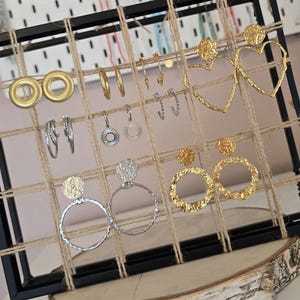 Peut inclure: Présentation de boucles d'oreilles dorées et argentées. Divers modèles sont exposés, dont des créoles, des clous et des pendants, avec des finitions texturées et lisses. L'ensemble est présenté sur un support grillagé avec une base en bois.