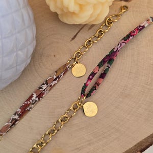 Peut inclure: Deux bracelets composés de chaînes en métal doré et de rubans en tissu à motifs floraux. L'un des bracelets comporte une breloque ronde dorée avec un motif de soleil, l'autre une breloque ronde dorée gravée du prénom Lola en écriture cursive.