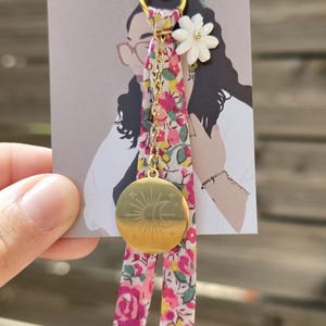 Peut inclure: Un porte-clés doré avec un ruban floral et un pendentif rond représentant un soleil et une lune. Le porte-clés comprend une fleur blanche et une chaîne dorée. La carte derrière le porte-clés porte l'inscription "Ptit Gravure bijoux".