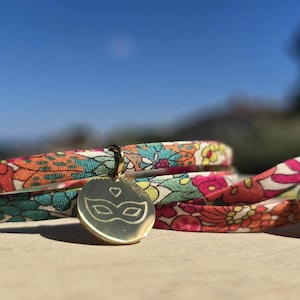 Peut inclure: Deux bracelets en tissu arborant un motif floral aux tons orange, bleu sarcelle, rose et jaune. Chaque bracelet est orné d'une breloque ronde en métal doré poli, gravée d'un petit cœur et d'un motif de masque de carnaval.