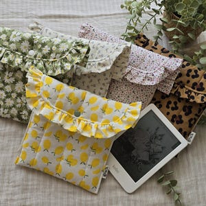Peut inclure: Une collection de housses matelassées en tissu pour liseuses, chacune dotée d'un rabat à volants avec fermeture à pression. Les motifs incluent des marguerites blanches sur fond vert, des petites fleurs et un imprimé léopard. Une housse aux citrons contient une Kobo.