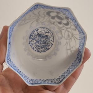 Puede incluir: Un cuenco octogonal de porcelana blanca con un borde floral azul y un diseño central de dragón azul. El cuenco tiene un patrón delicado y adornado y se sostiene en una mano.