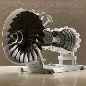 May include: A white 3D printed model of a jet engine with a cutaway view of the internal components. The engine is mounted on a white stand.