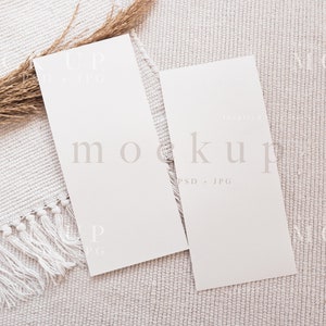 May include: Two blank white cards on a cream-colored textured surface with a dried grass stem. The cards are vertical and have a subtle texture. The text "mockup PSD + JPG" is printed in light gray on the right card.