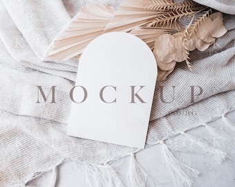 Può includere: Un cartoncino bianco con bordi arrotondati e una parte superiore leggermente arcuata, appoggiato su un tessuto bianco testurizzato con una trama naturale intrecciata. Il cartoncino presenta la parola "MOCKUP" stampata in un font marrone chiaro. Il tessuto è parzialmente ricoperto da materiale vegetale naturale essiccato, tra cui una foglia di palma secca a forma di ventaglio.