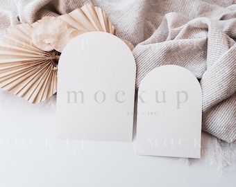 Può includere: Due carte bianche a forma di arco con la parola "mockup" stampata su di esse, appoggiate su una superficie bianca con una coperta di tessuto beige e un ventaglio di carta beige.