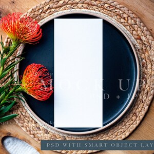 May include: A blank white card mockup on a black plate with a woven placemat. The plate is on a wooden surface with a green plant with orange flowers.