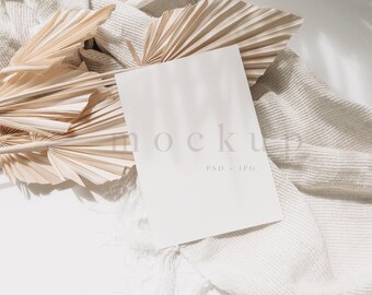 Puede incluir: Una tarjeta blanca en blanco con la palabra "mockup" impresa, apoyada sobre una tela beige texturizada. Hojas de palma secas están dispuestas alrededor de la tarjeta. El texto "PSD + JPG" está impreso debajo de la palabra "mockup".