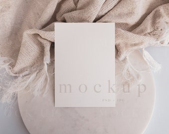 Puede incluir: Una maqueta de tarjeta blanca sobre una superficie de mármol con una tela beige texturizada drapeada sobre ella. La tarjeta tiene la palabra "mockup" impresa en ella en una fuente gris claro.