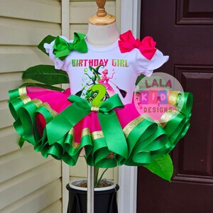 May include: A white t-shirt with the text "BIRTHDAY GIRL" and a graphic of two witches, paired with a pink tutu skirt trimmed with green and gold ribbon. The outfit includes green and pink bows and is displayed on a mannequin.