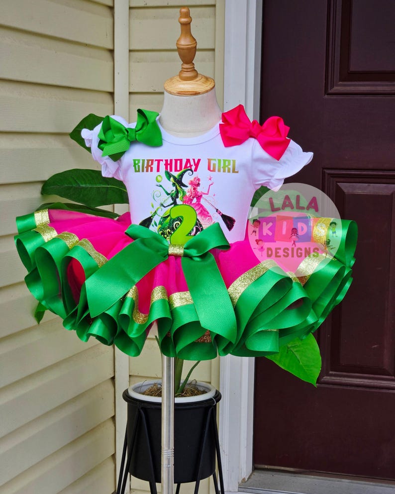 May include: A white t-shirt with the text "BIRTHDAY GIRL" and a graphic of two witches, paired with a pink tutu skirt trimmed with green and gold ribbon. The outfit includes green and pink bows and is displayed on a mannequin.