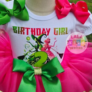 May include: A birthday outfit featuring a white t-shirt with the words "BIRTHDAY GIRL" and a graphic of two witches. The outfit includes a pink tutu skirt, green bows, and a gold glitter accent. The number "2" is prominently displayed.