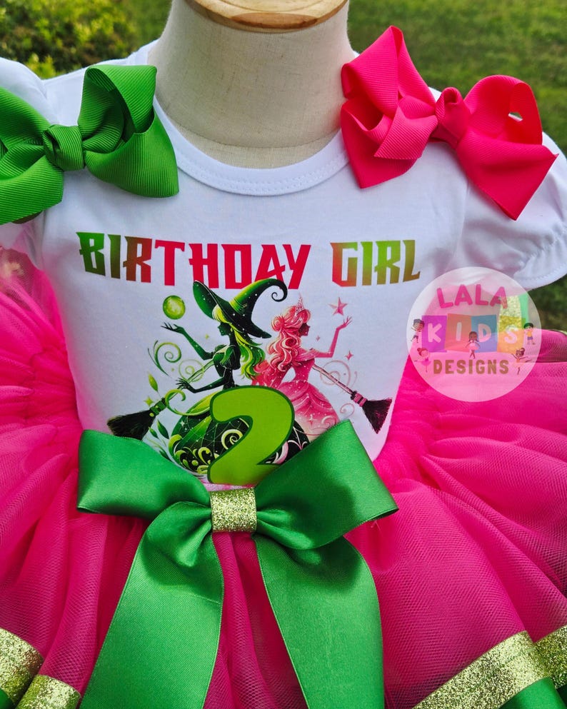 May include: A birthday outfit featuring a white t-shirt with the words "BIRTHDAY GIRL" and a graphic of two witches. The outfit includes a pink tutu skirt, green bows, and a gold glitter accent. The number "2" is prominently displayed.