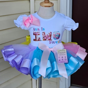 May include: A white t-shirt with the text "Ava is TWO Sweet" and a colorful tutu. The tutu is layered with purple, blue, and pink tulle, accented with matching satin ribbons. A large pink bow adorns the top.