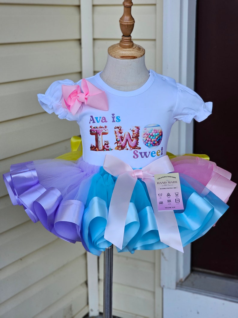 May include: A white t-shirt with the text "Ava is TWO Sweet" and a colorful tutu. The tutu is layered with purple, blue, and pink tulle, accented with matching satin ribbons. A large pink bow adorns the top.
