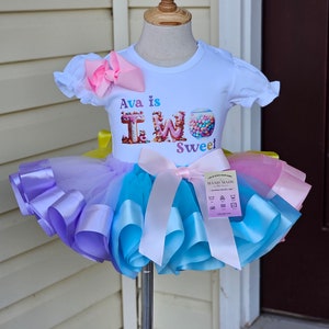 May include: A white t-shirt with the text "Ava is TWO Sweet" and a colorful tutu. The tutu is a mix of purple, blue, yellow, and pink. A large pink bow is attached to the shirt, and a smaller bow adorns the tutu.