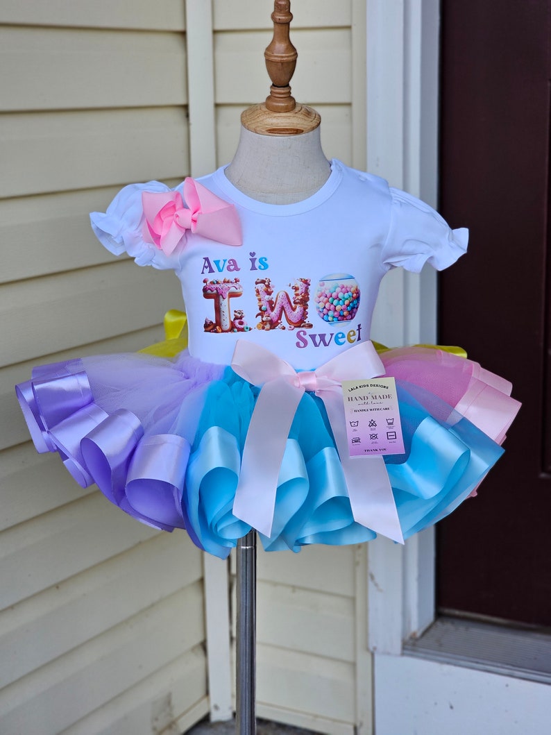 May include: A white t-shirt with the text "Ava is TWO Sweet" and a colorful tutu. The tutu is a mix of purple, blue, yellow, and pink. A large pink bow is attached to the shirt, and a smaller bow adorns the tutu.
