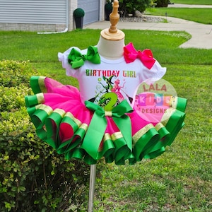 May include: A birthday outfit for a young child. The outfit includes a white t-shirt with the words "BIRTHDAY GIRL" and a graphic. The tutu is pink with green and gold ribbon trim. Green and pink bows complete the look.