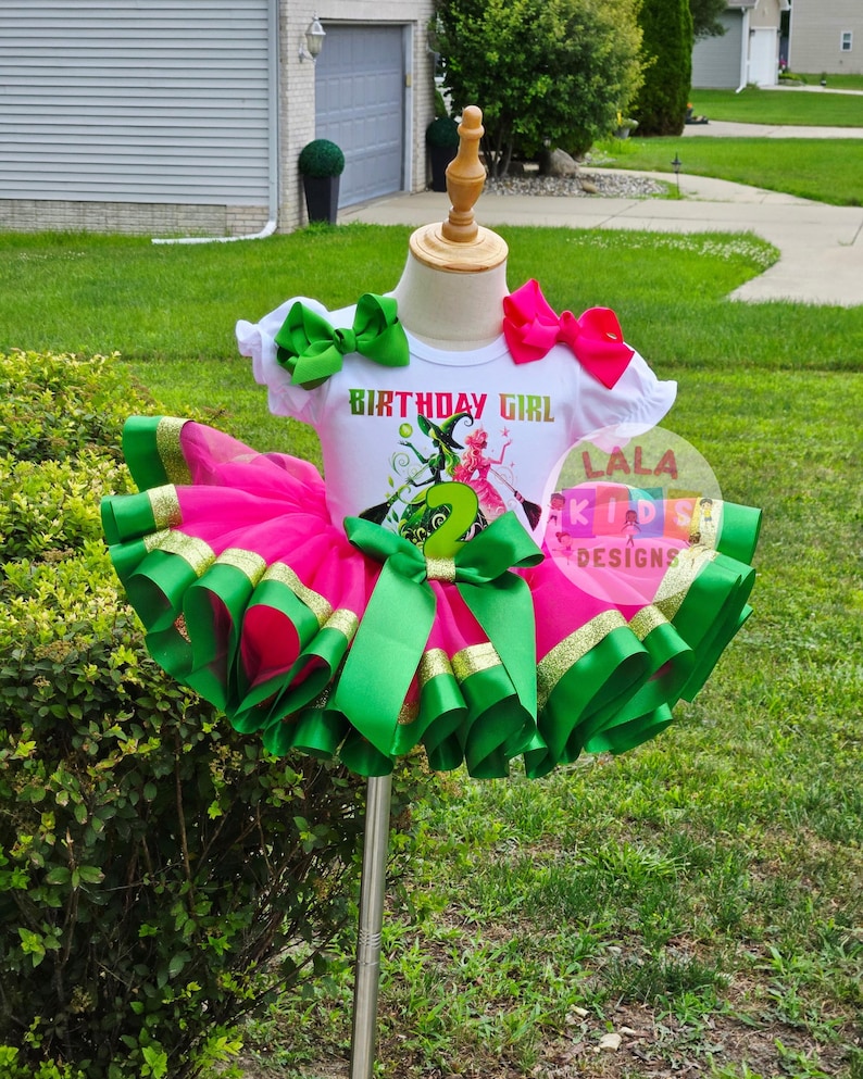 May include: A birthday outfit for a young child. The outfit includes a white t-shirt with the words "BIRTHDAY GIRL" and a graphic. The tutu is pink with green and gold ribbon trim. Green and pink bows complete the look.