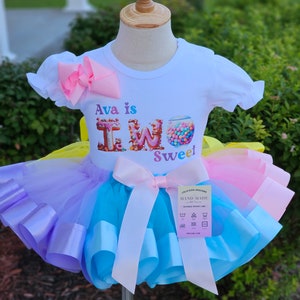 May include: A white t-shirt with the text "Ava is TWO Sweet" and a colorful candy graphic, paired with a multi-colored tutu skirt. The tutu features layers of blue, purple, yellow, and pink tulle with matching ribbon accents and a large pink bow.