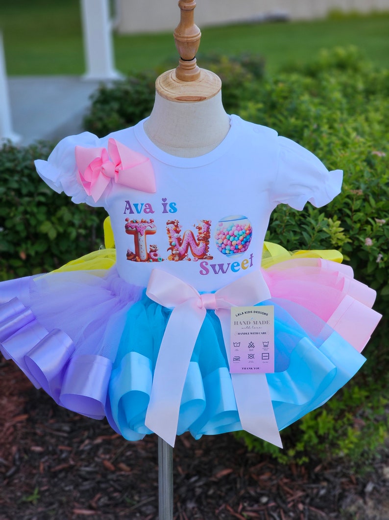 May include: A white t-shirt with the text "Ava is TWO Sweet" and a colorful candy graphic, paired with a multi-colored tutu skirt. The tutu features layers of blue, purple, yellow, and pink tulle with matching ribbon accents and a large pink bow.