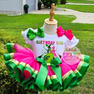 May include: A festive birthday outfit for a child, featuring a white t-shirt with the text "BIRTHDAY GIRL" and a graphic of a witch and a fairy. The outfit includes a pink tutu skirt with green and gold ribbon trim, and green bows.