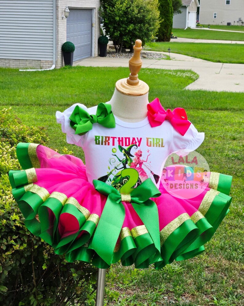 May include: A festive birthday outfit for a child, featuring a white t-shirt with the text "BIRTHDAY GIRL" and a graphic of a witch and a fairy. The outfit includes a pink tutu skirt with green and gold ribbon trim, and green bows.