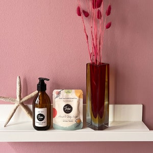 Puede incluir: Un estante blanco con una botella de vidrio marrón de lavado de manos y cuerpo con un dispensador de bomba negro. La botella tiene una etiqueta que dice "Trew" y "Hand & Body Wash". Hay una pequeña bolsa rosa y blanca con el texto "Trew" y "Hand & Body Wash" en ella. Un jarrón de vidrio rojo con flores secas rosas está en el estante.