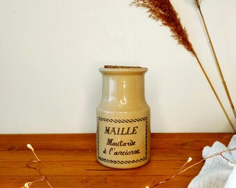 May include: A ceramic jar with a cork lid and a label that reads "MAILLE Mustard à l'ancienne". The jar is beige with brown lettering and is sitting on a wooden surface.